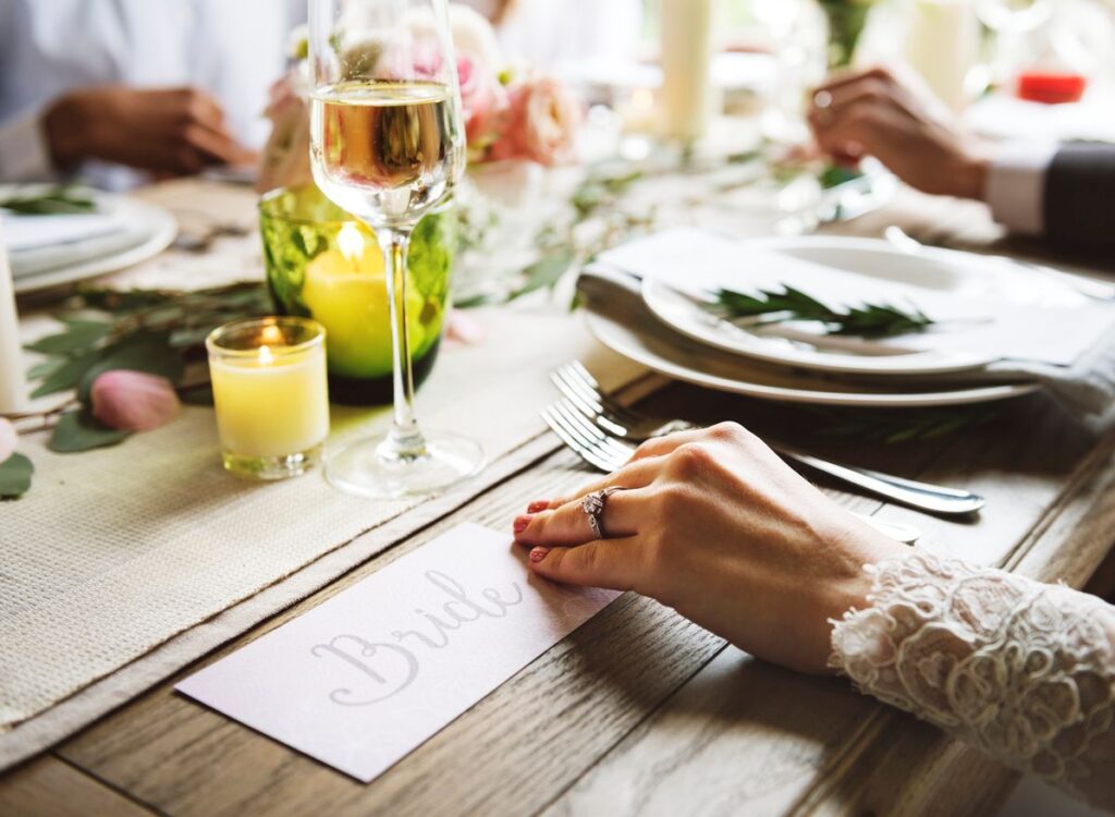 A bride’s hand with a wedding ring rests on a name card at a decorated reception table with candles. 