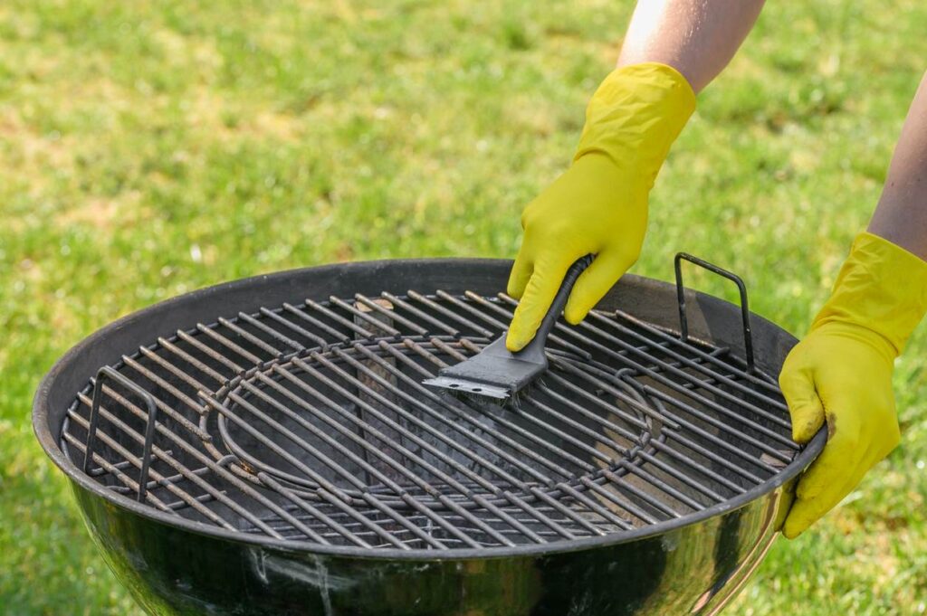  A person wearing yellow gloves cleans a round barbecue grill grate with a scraper tool in a sunny backyard. 