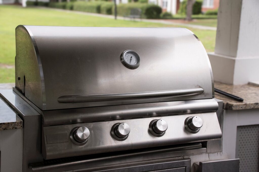 A closed stainless steel gas grill with four control knobs and a built-in thermometer on a covered patio near a lawn.