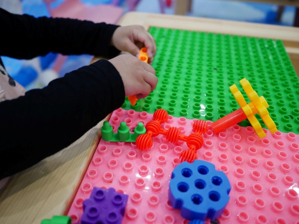 A child’s hands building with colorful interlocking toys on a green and pink pegboard at a play table.