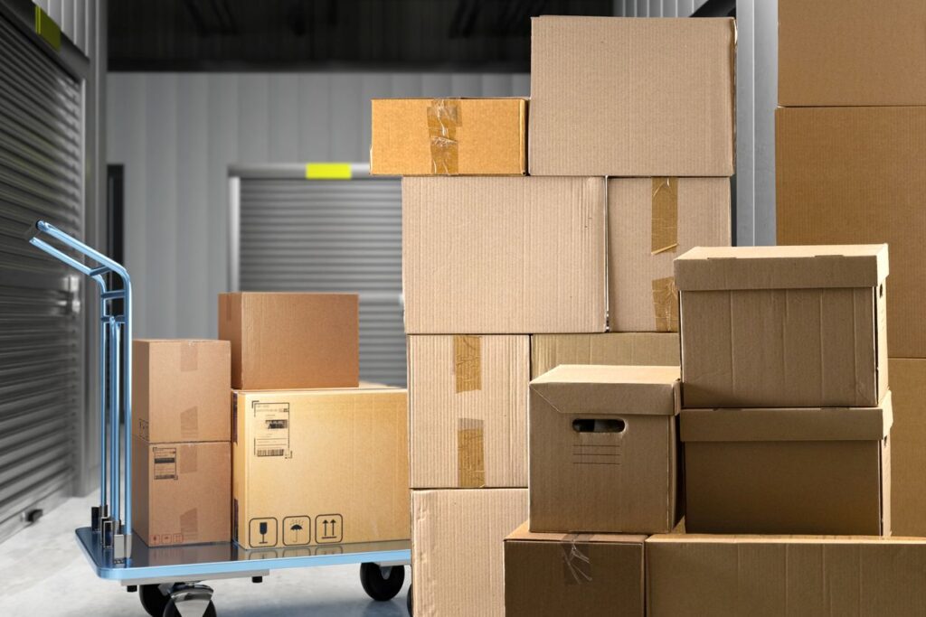 Stacked cardboard boxes and a moving cart inside a storage facility hallway with closed metal unit doors. 